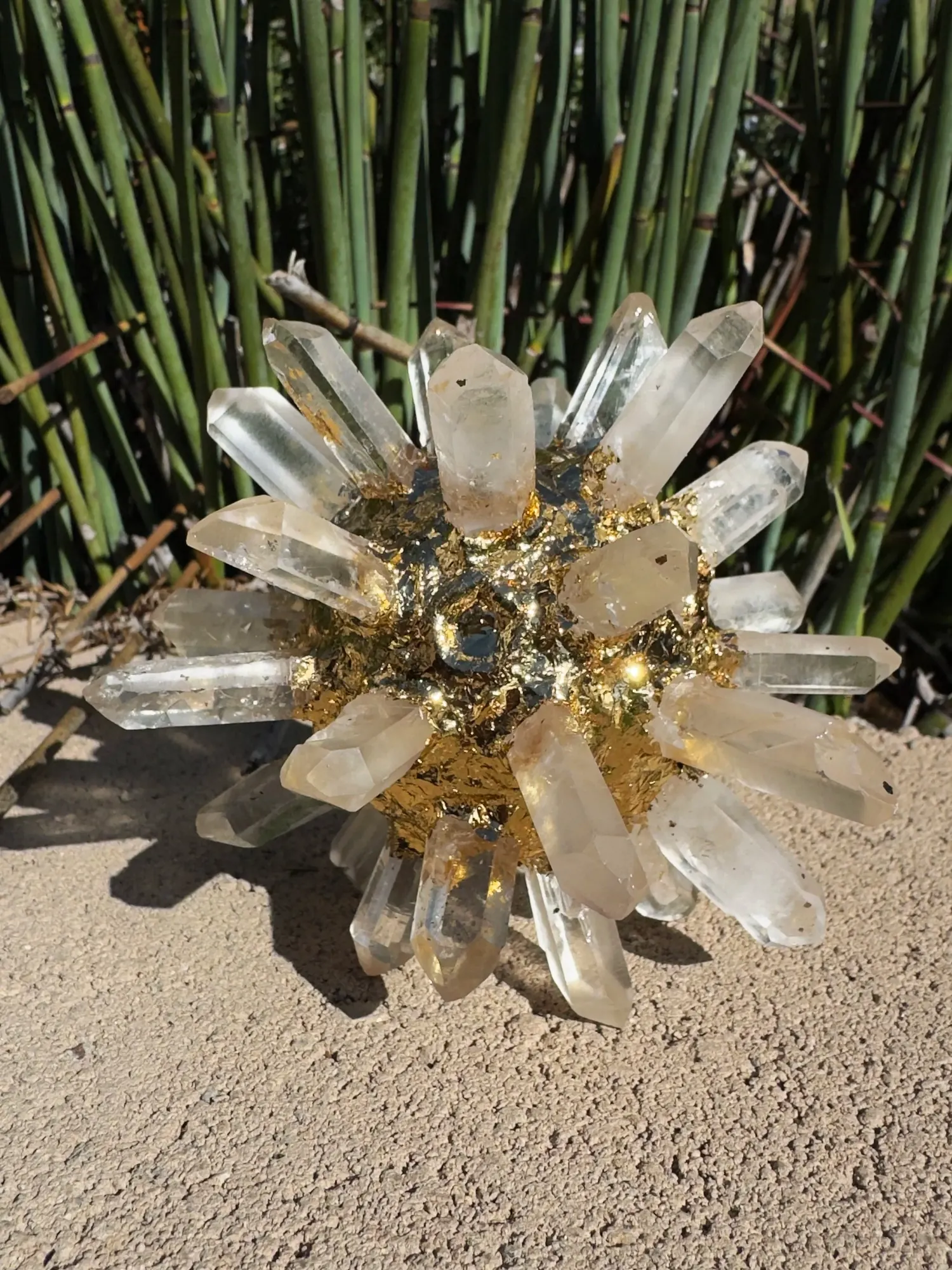 Lemurian Quartz Crystal AllSphyr: clear quartz points radiating from a gold-gilded core, on sandy ground in front of green stalks