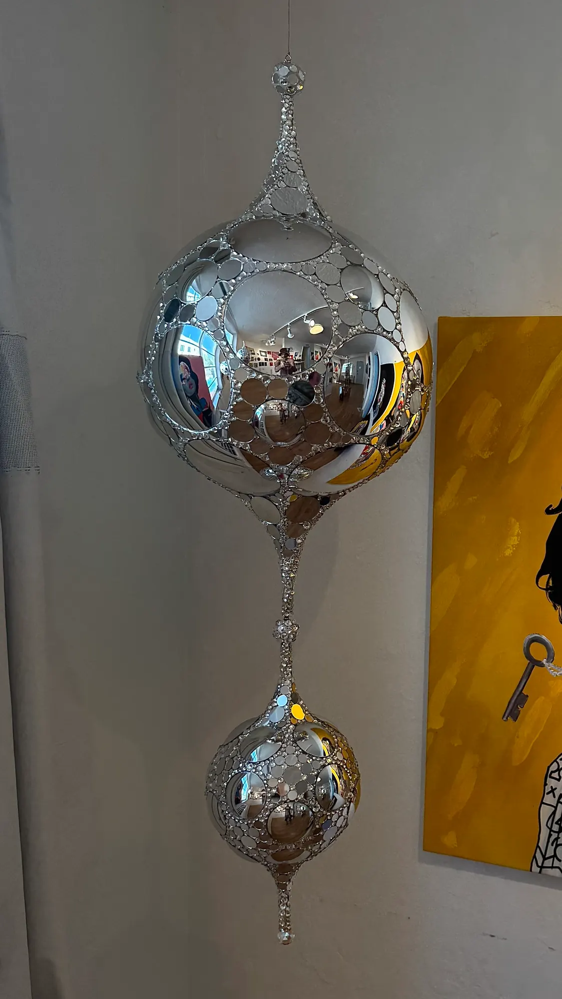 Teardrop DiscoSphyr suspended indoors beside a yellow painting, showing its large mirrored sphere and smaller lower drop