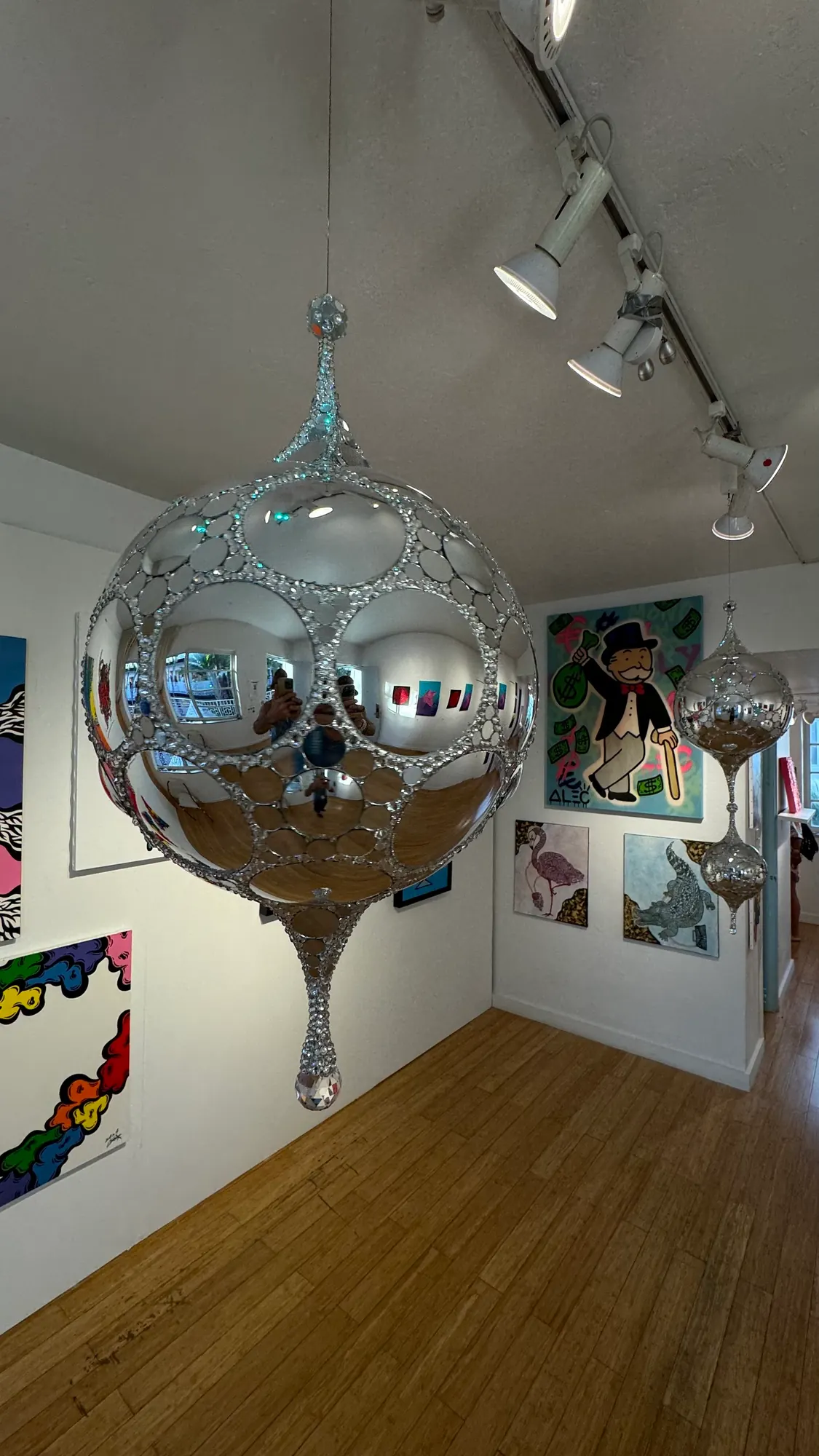 Galactic DiscoSphyr: a mirrored sphere with a rhinestone lattice and a crystal drip, suspended from a gallery ceiling among framed artworks